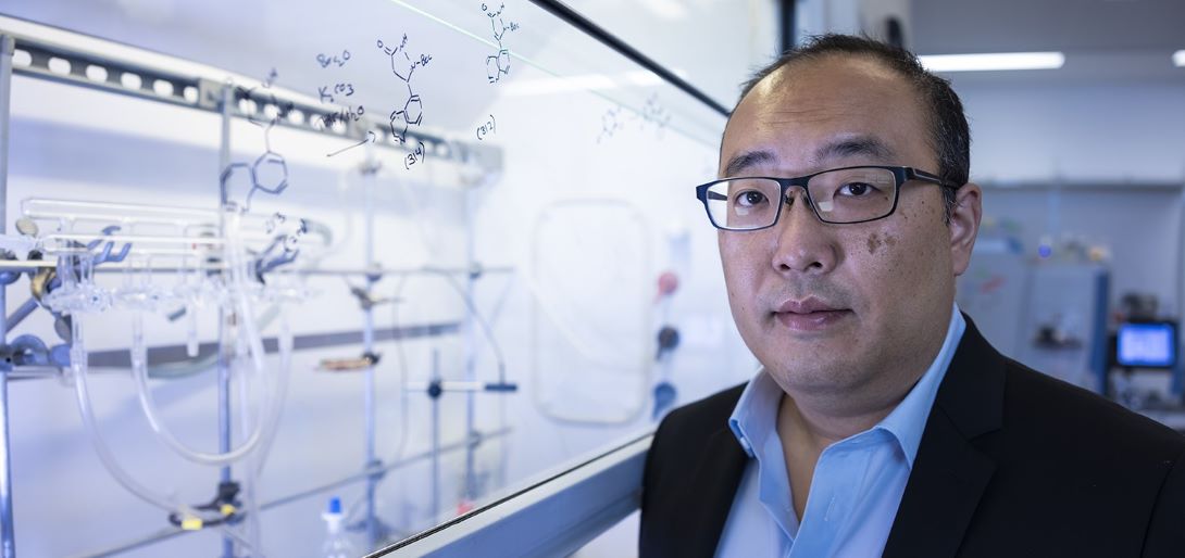 Dr. Daniel K. Nomura stands in front of a chemical fume hood in a chemistry laboratory wearing a light blue button-down shirt, a black blazer, and black glasses. There are chemical structures and reactions drawn on the fume hood sash.